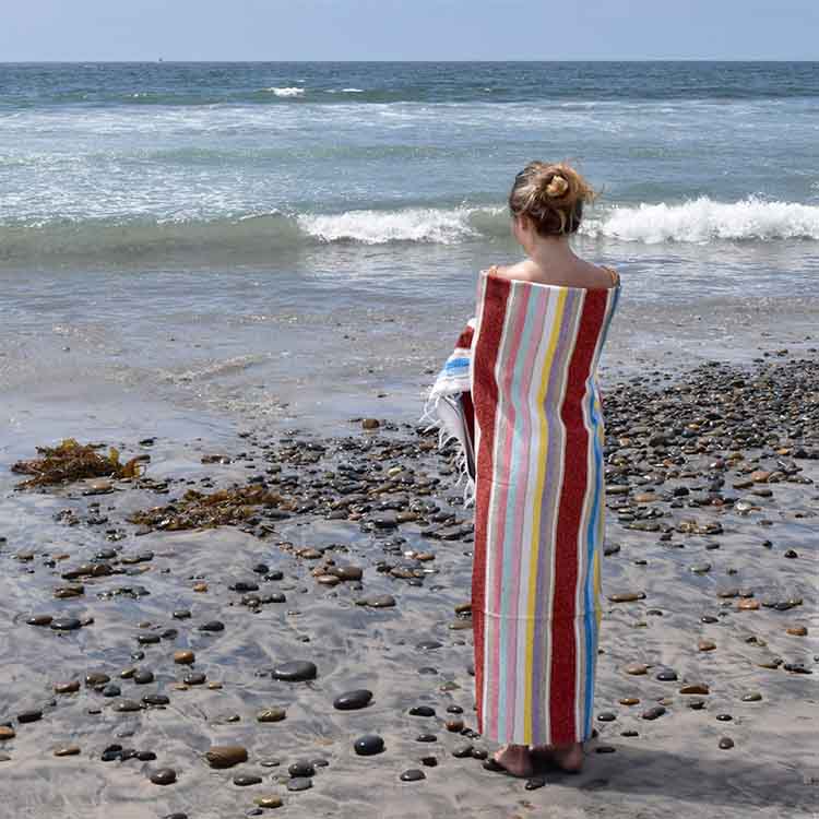Person wrapped in a striped towel on a pebbly beach with ocean waves in the background