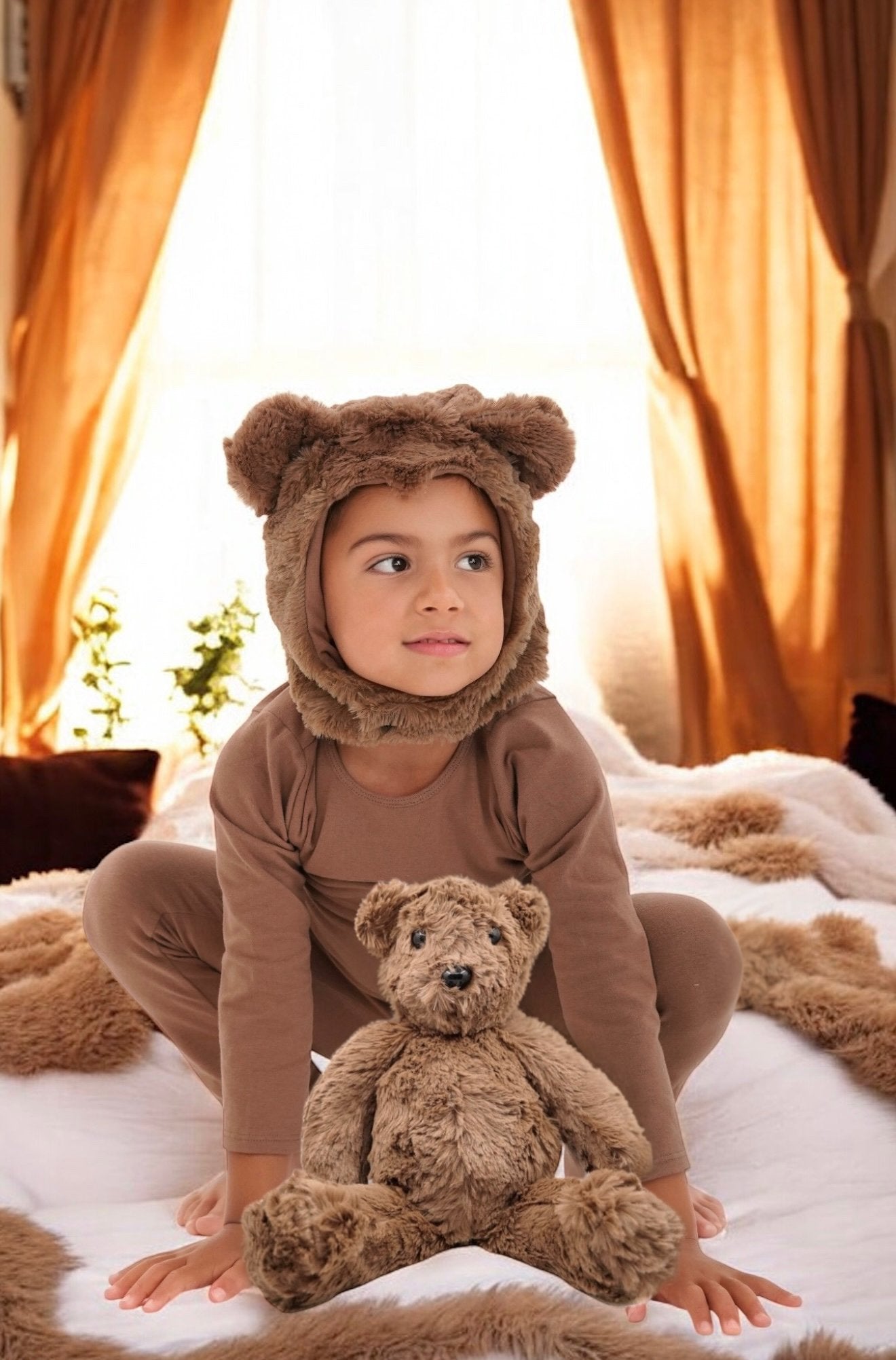 Child in a bear costume sitting on a bed with a teddy bear, surrounded by orange curtains.