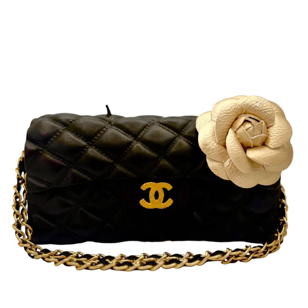 Black quilted handbag with gold chain and beige flower accessory on a gray background
