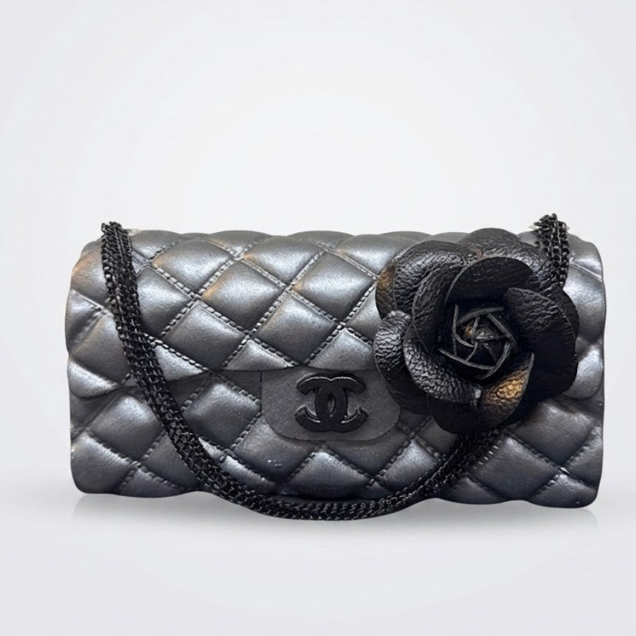 A black quilted leather shoulder bag with a large black camellia flower embellishment and a black chain strap, shown against a light gray background.