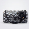 A black quilted leather shoulder bag with a large black camellia flower embellishment and a black chain strap, shown against a light gray background.