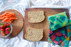 Two slices of bread on a wooden cutting board with a container of strawberries and a bowl of carrots, all on a white surface.