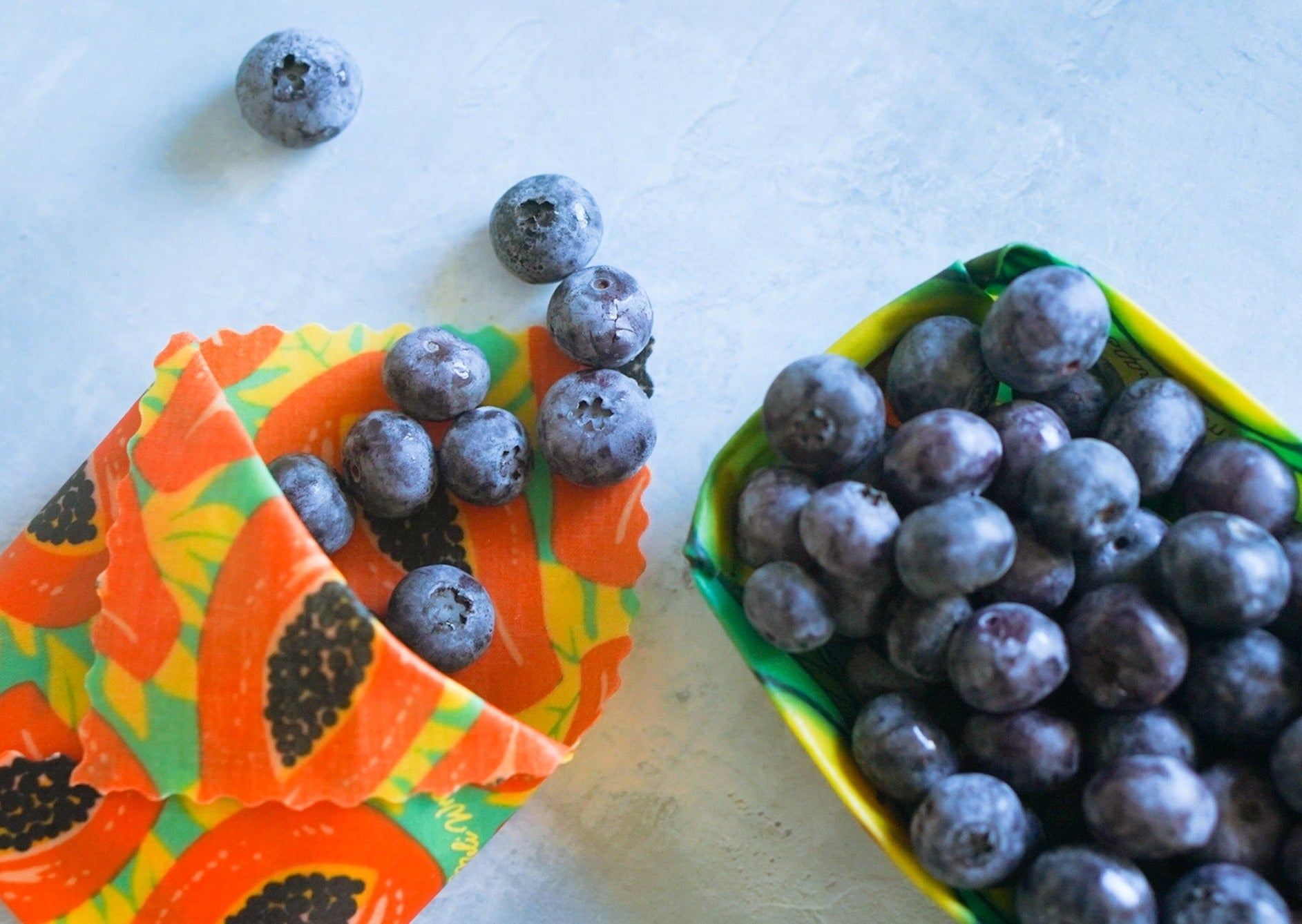 Blueberries on a colorful paper plate and in a green container on a light surface.