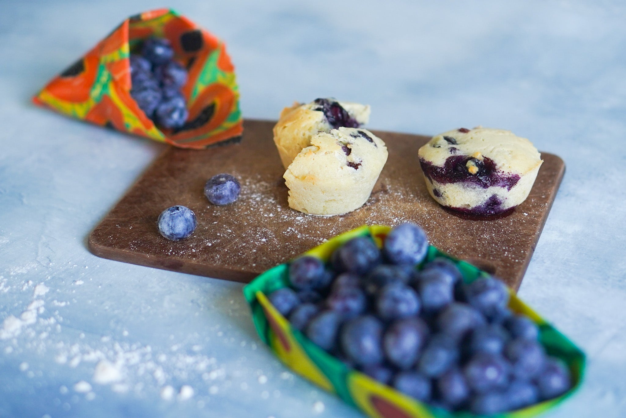 Blueberry muffins on a wooden board with blueberries and colorful packaging in the background.
