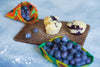 Blueberry muffins on a wooden board with blueberries and colorful packaging in the background.