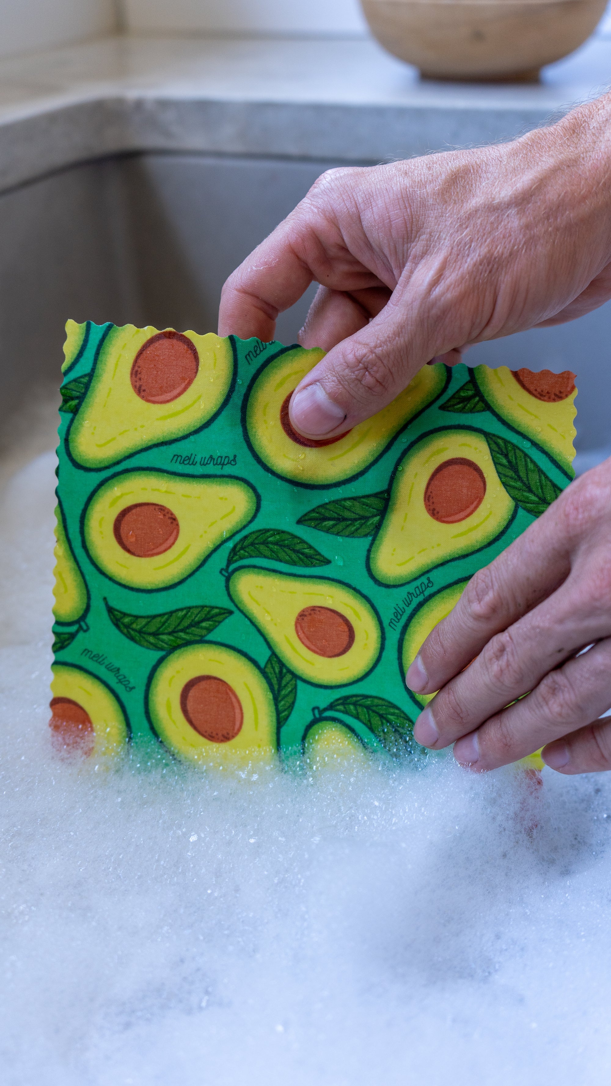 Hand holding a colorful avocado-patterned sponge over a sink with soapy water.