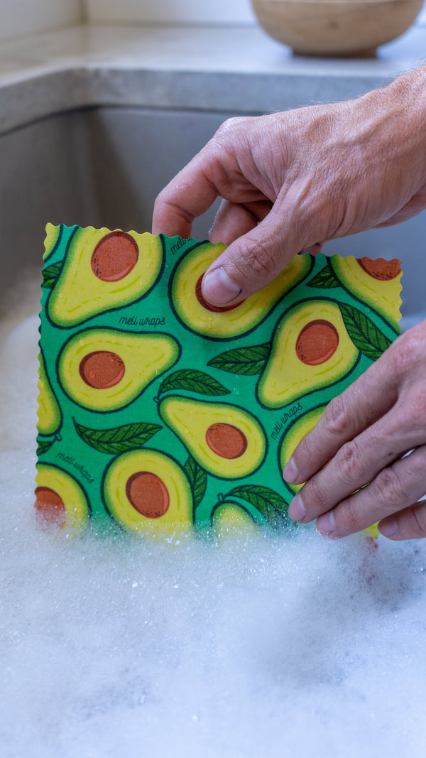 Hand holding a colorful avocado-patterned sponge over a sink with soapy water.