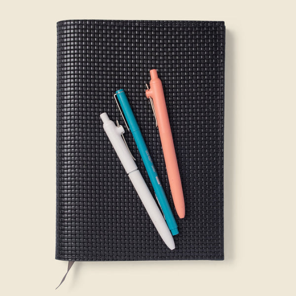 Black woven notebook with three pens on a beige background