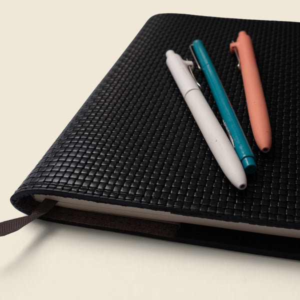 Black textured notebook with three pens on a light beige background