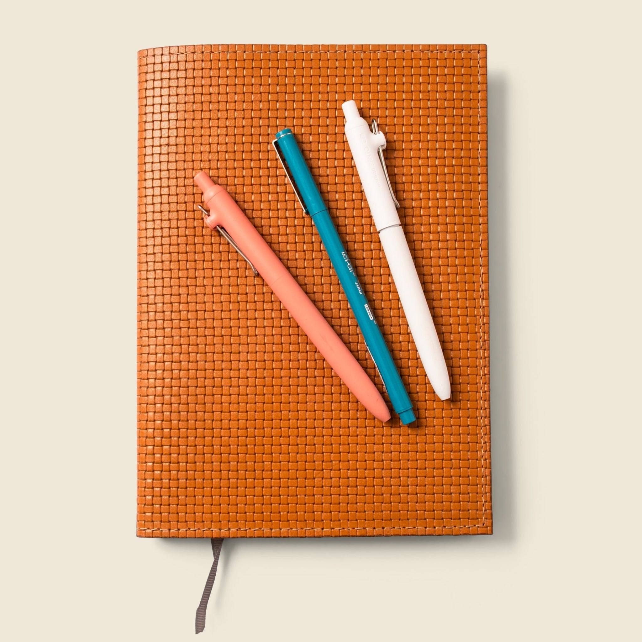 Orange woven notebook with three pens on a beige background
