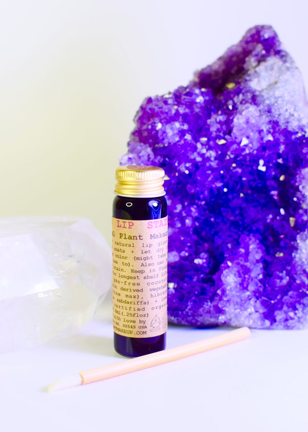 Lip balm bottle with purple label next to a large amethyst crystal on a white background