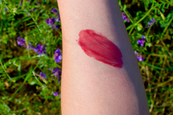 Pink lipstick swatch on a person's arm with a natural background of greenery and flowers.