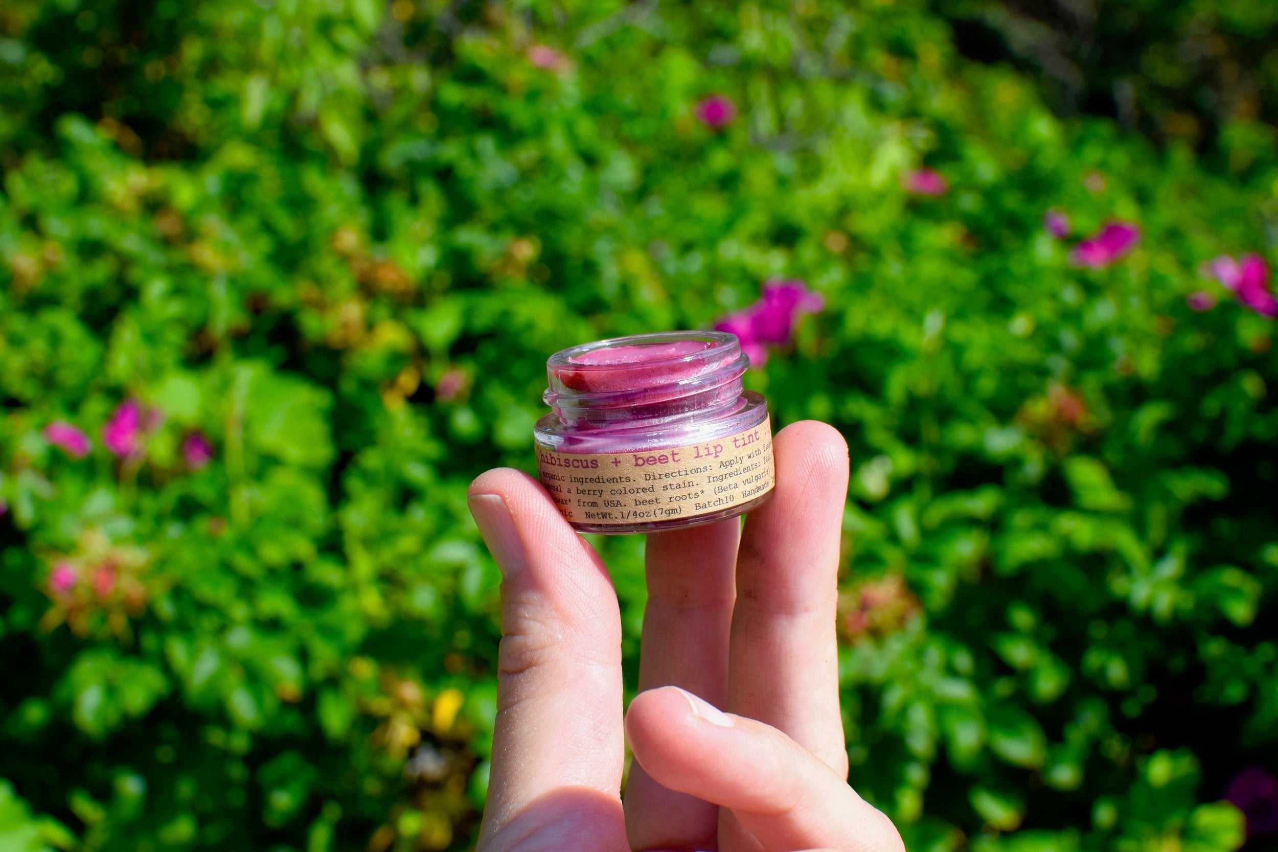 Hand holding a small jar of lip balm against a green outdoor background with flowers.