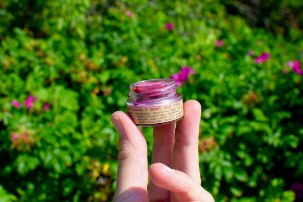 Hand holding a small jar of lip balm against a green outdoor background with flowers.