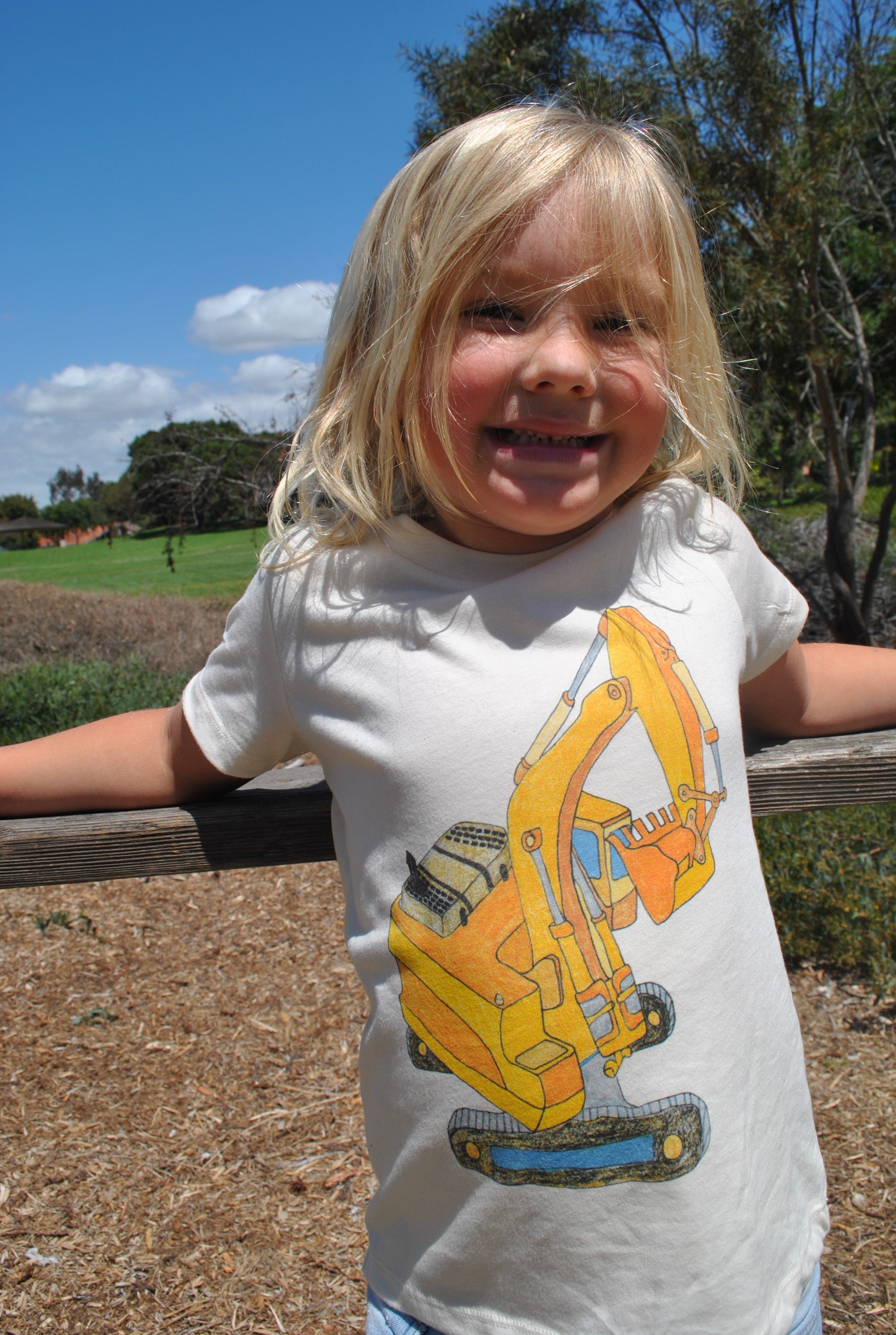 Child wearing a white t-shirt with a yellow excavator graphic outdoors.