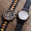 Two watches on a wooden surface, one with a wooden strap and detailed face, the other with a black strap and engraved message.