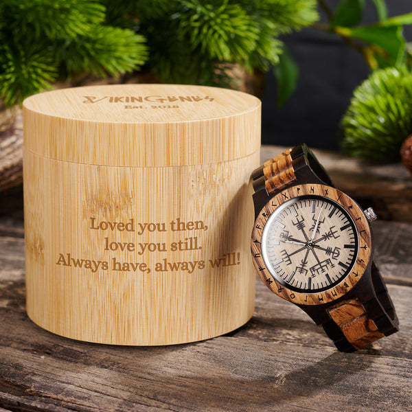 Wooden watch with engraved message on a wooden box, surrounded by plants.