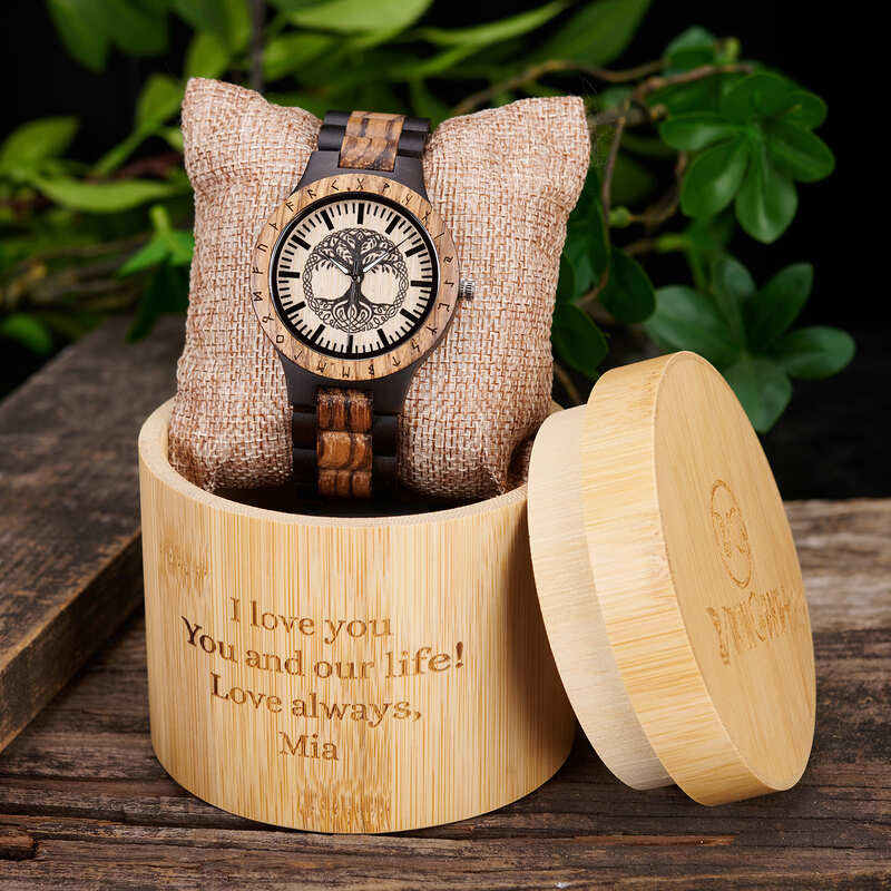 Wooden watch with tree of life design in a bamboo box with engraved message, surrounded by greenery.