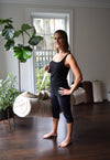 Woman in black tank top and leggings standing in a room with plants and a window.