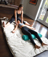 Two women in a home setting, one lying on a mat with a green cylindrical object, the other assisting.