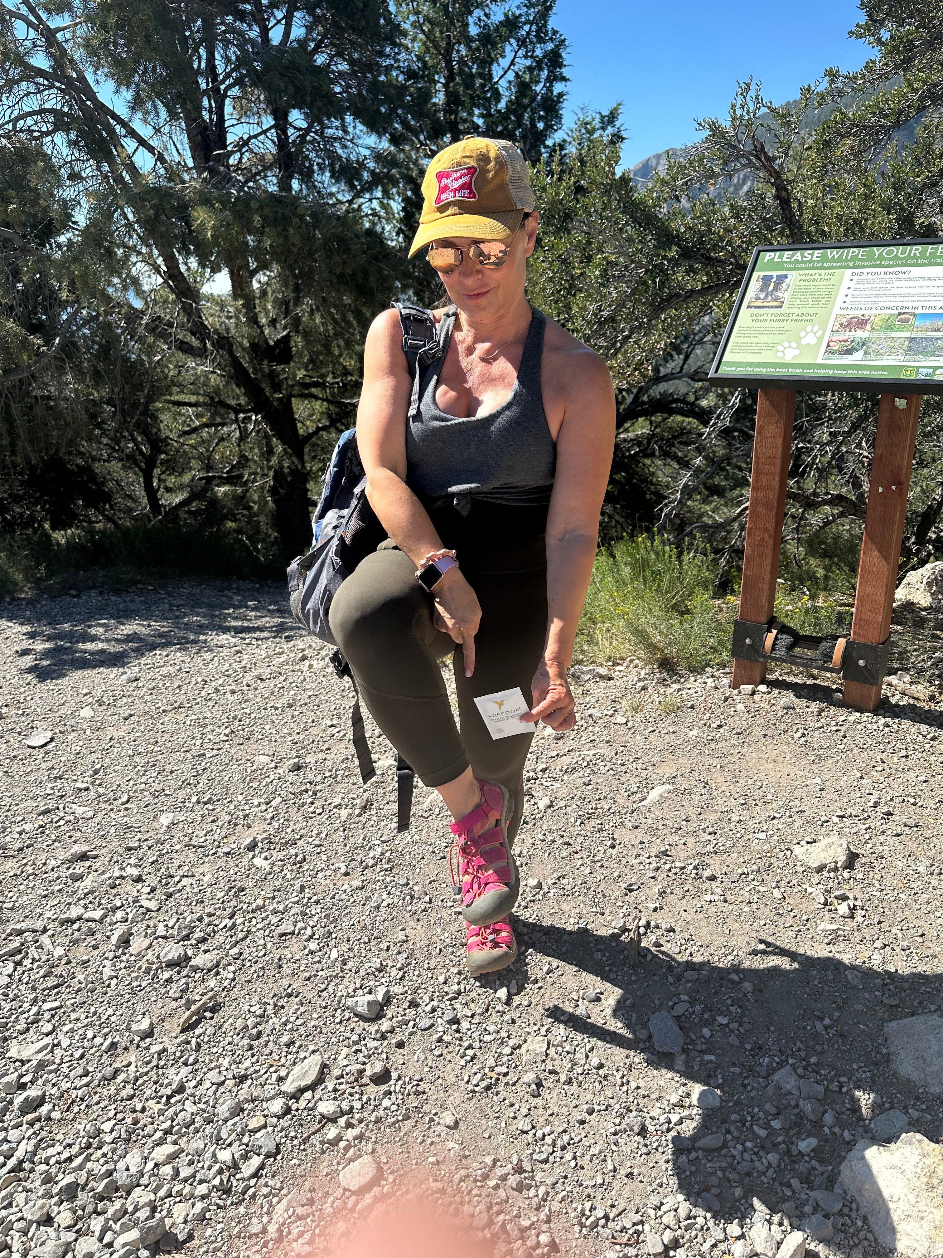 Woman hiking on a trail with a backpack and sunglasses, standing next to a informational sign.