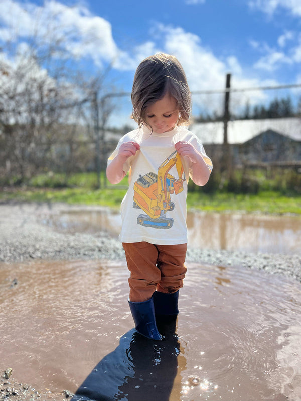 Child in a white t-shirt with a truck design and blue rain boots standing in a puddle outdoors.