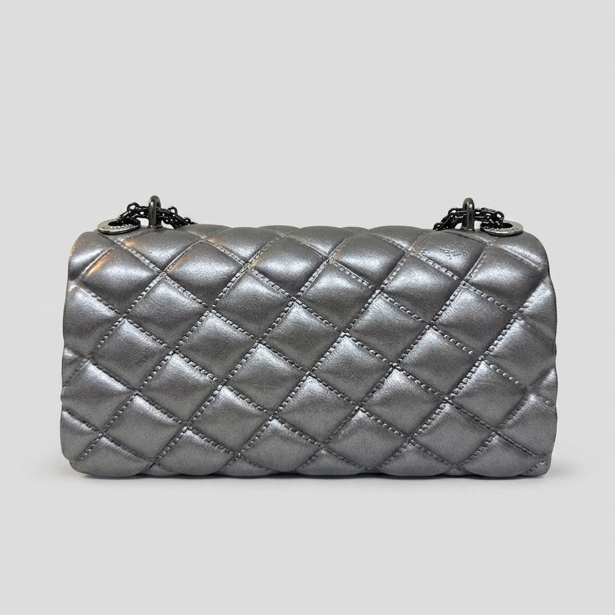 A metallic silver quilted shoulder bag with a diamond pattern and a chain strap, shown against a solid light gray background.