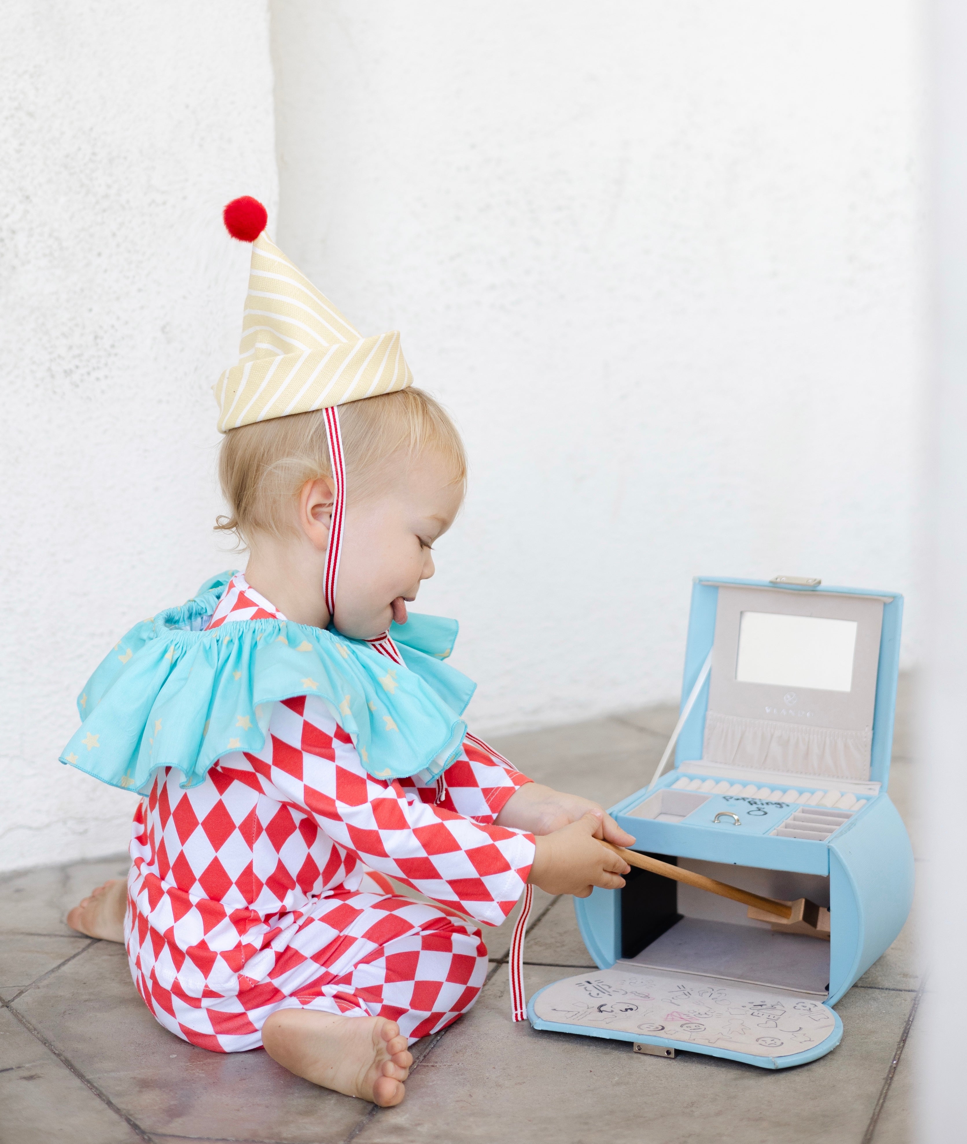Child in clown costume playing with a toy cash register against a white wall.
