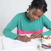 Young girl in a teal and pink shirt coloring with crayons on a white sheet of paper.
