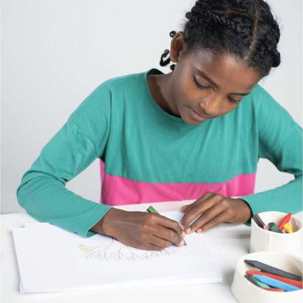 Young girl in a teal and pink shirt coloring with crayons on a white sheet of paper.