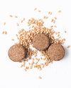 Three round brown cookies surrounded by scattered whole grains on a white background