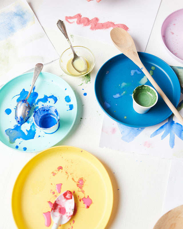 Colorful plates with paint splashes and utensils on a white surface