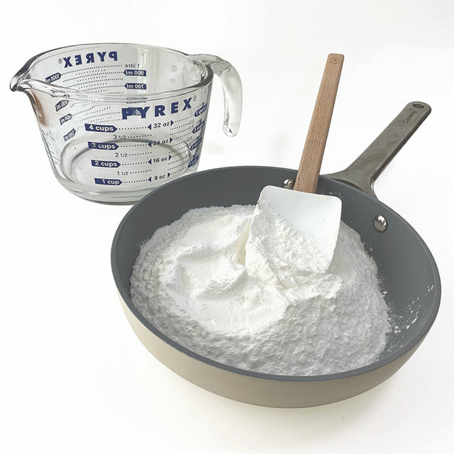 Pyrex measuring cup next to a pan with flour and a spatula on a white background
