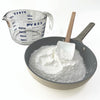 Pyrex measuring cup next to a pan with flour and a spatula on a white background