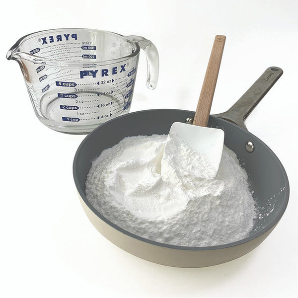 Pyrex measuring cup next to a pan with flour and a spatula on a white background
