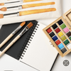 Watercolor travel bundle contents laid out including bamboo watercolor case, brushes, and watercolor pad
