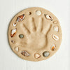 Round sand mold with a handprint and seashells on a white background