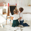 Two children drawing on a blackboard easel in a bright living room.