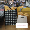 Two gift bags with yellow ribbons on a wooden surface with a blurred Christmas tree in the background.