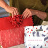Person wrapping a gift with red paper and a red bow, with a white card featuring blue and red patterns.