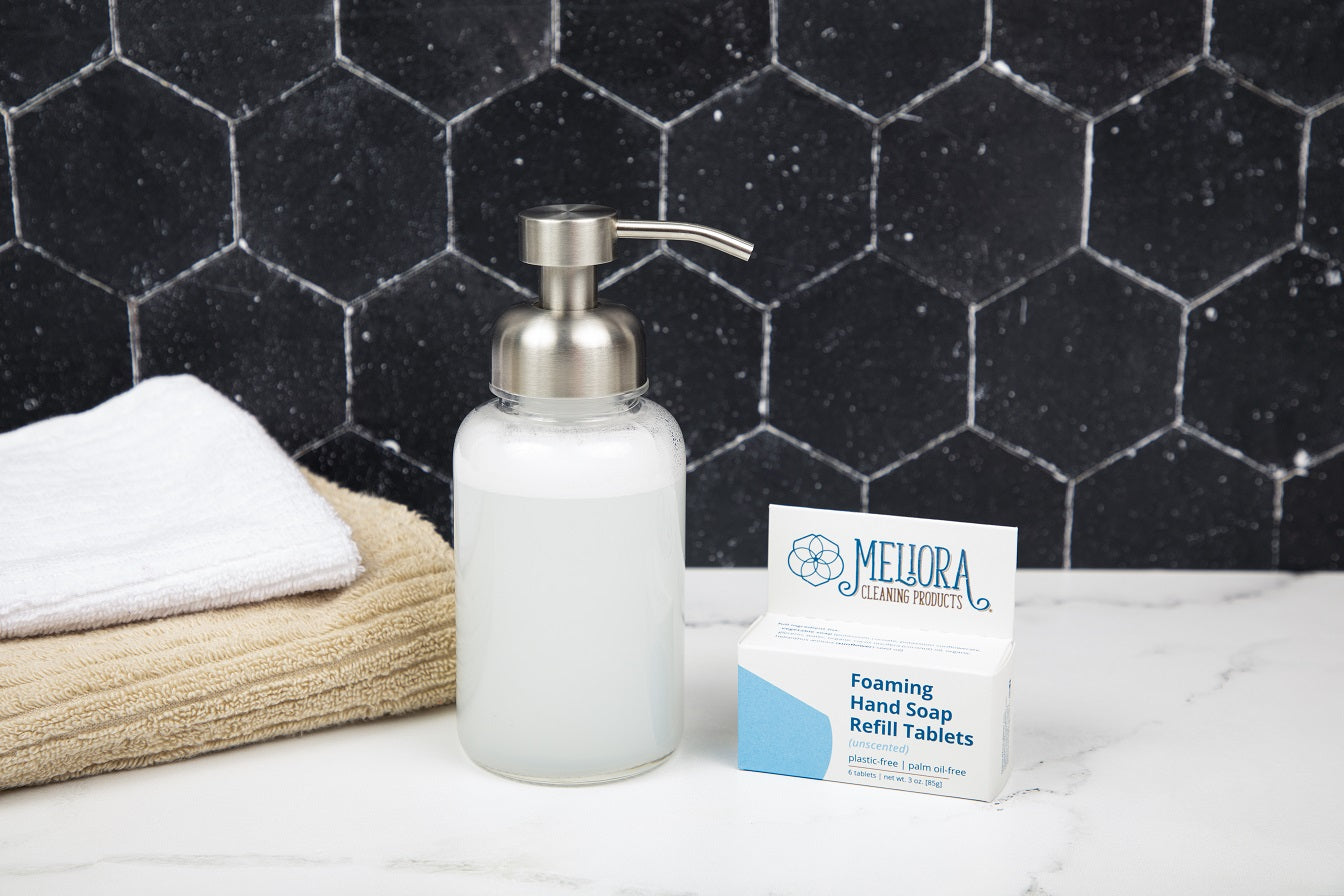 Foaming hand soap dispenser and refill tablets on a bathroom counter with a black tiled wall background.