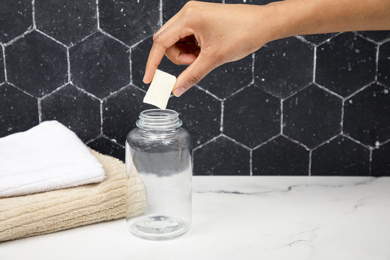 Person adding a white cube to a clear bottle on a marble surface with a black tiled wall background.