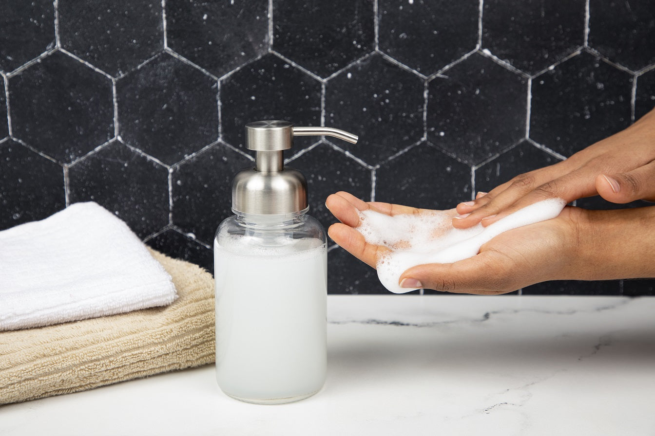 Hand soap dispenser with soapy hands against a black tiled wall.