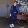 A child model wearing a navy blue tracksuit with a blue and white checkered jacket, posing next to a rustic wooden wagon in front of gray barn doors.