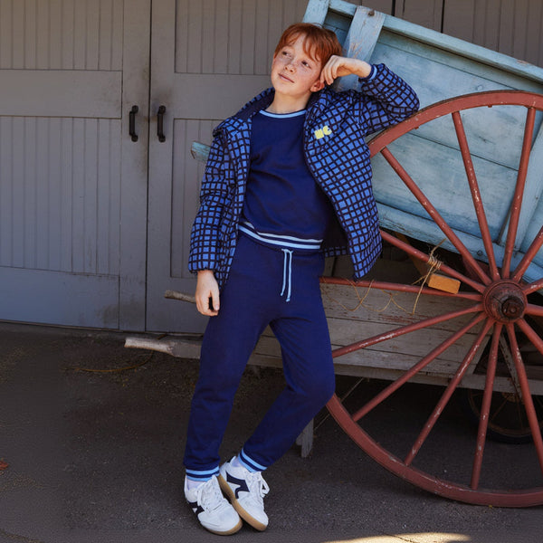 A child model wearing a navy blue tracksuit with a blue and white checkered jacket, posing next to a rustic wooden wagon in front of gray barn doors.