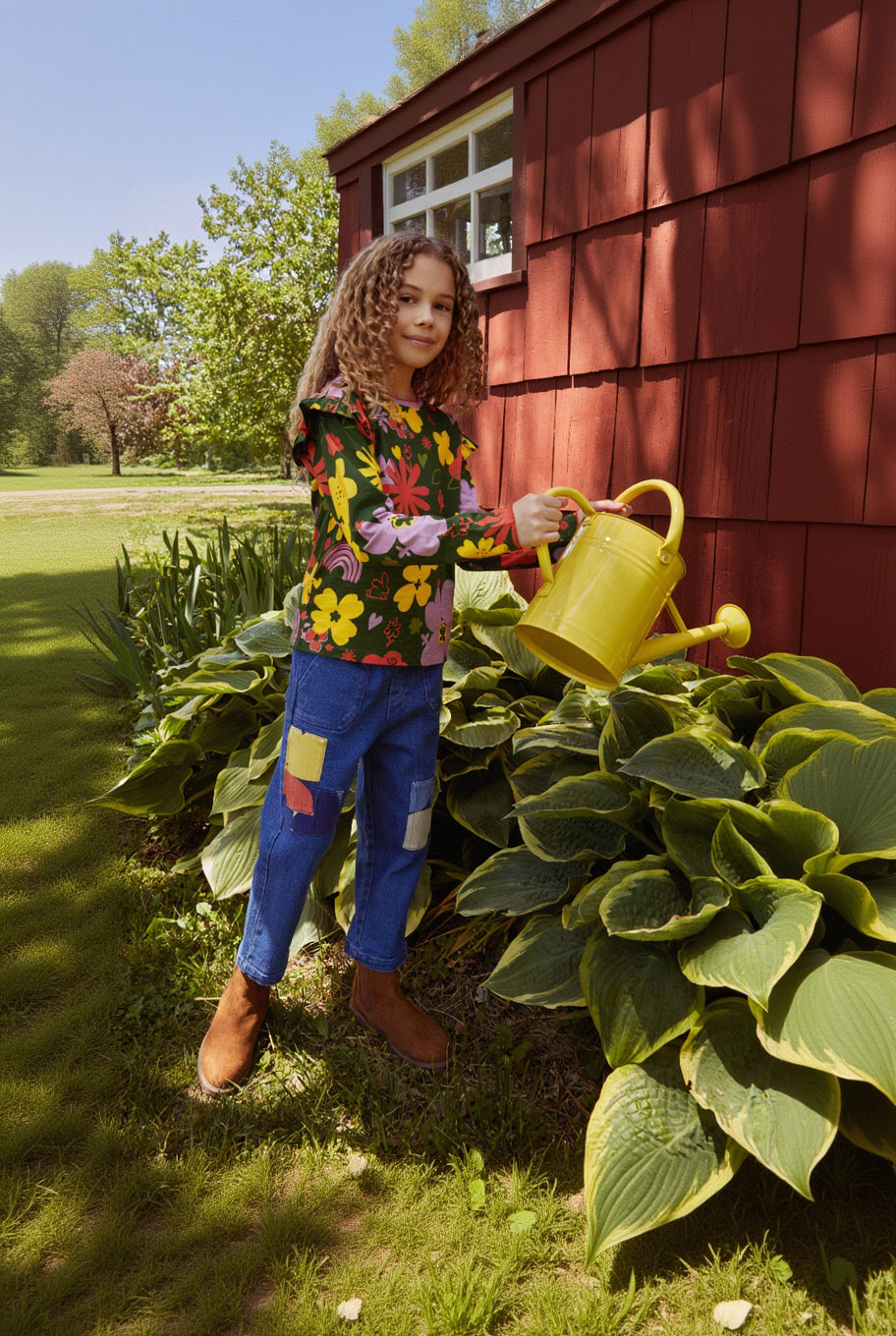 A woman stands in a garden next to a red wooden shed, holding a yellow watering can. She is wearing a colorful floral long-sleeve top, blue jeans with colorful patch details, and brown boots.
