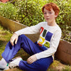 A young boy with red hair sits on grass in a garden setting, wearing a white long-sleeve graphic sweatshirt with a colorful geometric print and blue sweatpants.