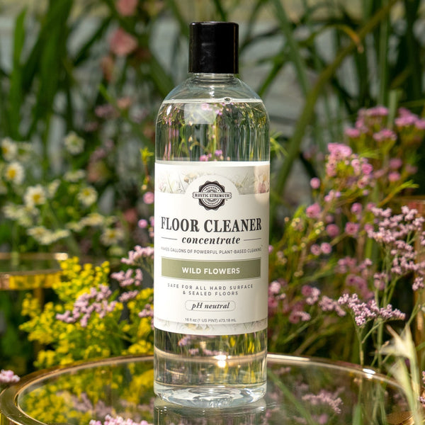 Floor cleaner bottle on a glass table with a natural background of plants and flowers. Wild Flowers floor cleaner concentrate — non-toxic, plant-based & pH neutral. Makes gallons, safe for kids & pets, 100% biodegradable & ammonia-free. Shop now.