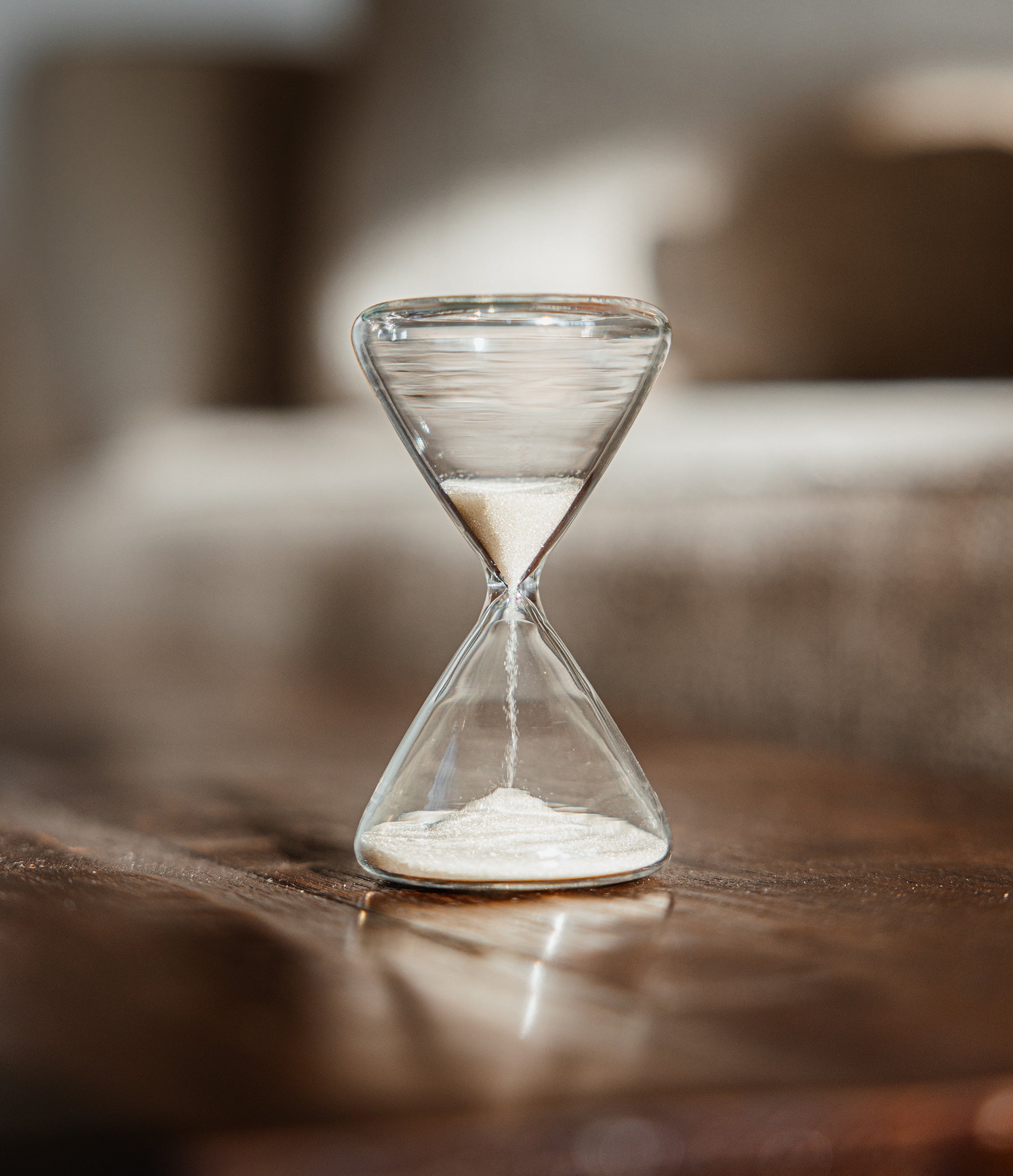 A clear glass hourglass filled with white sand, standing on a dark wooden surface with a softly blurred indoor background.