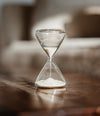 A clear glass hourglass filled with white sand, standing on a dark wooden surface with a softly blurred indoor background.
